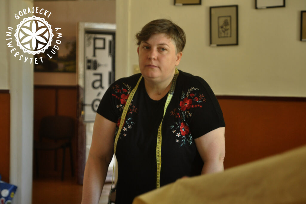 Kobieta w czarnej bluzce z czerwonym haftem, ma na szyi miarkę krawiecką, Miranda